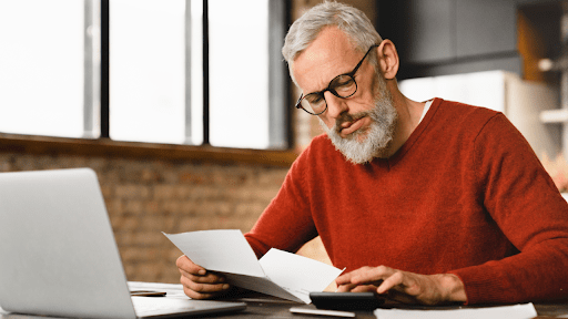 A senior man with glasses reviewing tax documents at a laptop, wondering when a tax debt expires