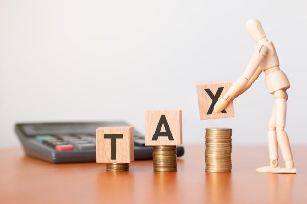 Wooden blocks spelling "TAX" on stacked coins with a calculator and mannequin figure—representing IRS debt and financial burden.