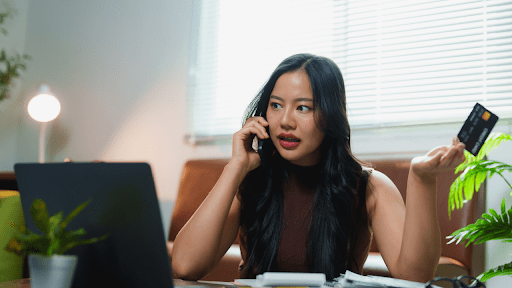 Young woman on phone holding credit card at laptop wondering why do I have a tax debt