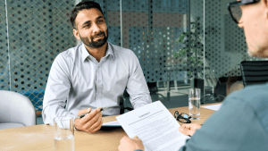This image shows a professional financial consultation taking place in a modern office setting, where a bearded man in a light gray dress shirt is meeting with a client to discuss important financial matters. The advisor appears to be explaining how tax obligations impact creditworthiness, likely addressing the common concern "does IRS debt affect credit" scores and lending decisions. A contract document is visible on the wooden conference table between them, suggesting they may be discussing how tax debt can influence loan applications, mortgage approvals, or other financial agreements. The contemporary office environment with glass partitions and professional atmosphere indicates this is a legitimate financial advisory meeting where clients seek guidance about whether IRS debt affect credit ratings and what steps they can take to protect their financial standing. The advisor's calm demeanor and the formal setting emphasize the importance of understanding how tax obligations intersect with credit reporting, as many taxpayers are concerned about does IRS debt affect credit scores when they're dealing with outstanding tax liabilities. This professional consultation represents the type of expert guidance needed to navigate the complex relationship between tax debt and credit health.