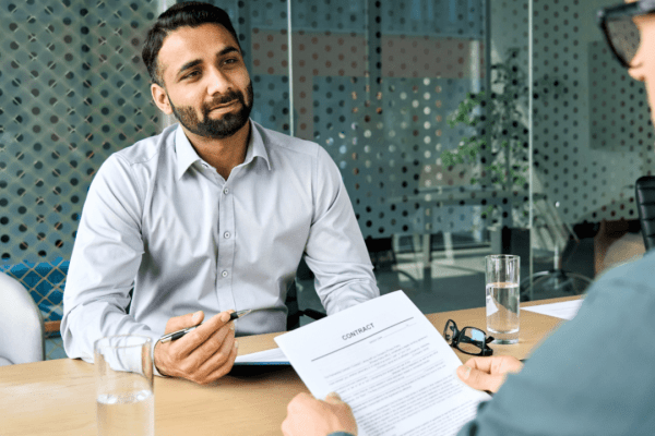 This image shows a professional financial consultation taking place in a modern office setting, where a bearded man in a light gray dress shirt is meeting with a client to discuss important financial matters. The advisor appears to be explaining how tax obligations impact creditworthiness, likely addressing the common concern "does IRS debt affect credit" scores and lending decisions. A contract document is visible on the wooden conference table between them, suggesting they may be discussing how tax debt can influence loan applications, mortgage approvals, or other financial agreements. The contemporary office environment with glass partitions and professional atmosphere indicates this is a legitimate financial advisory meeting where clients seek guidance about whether IRS debt affect credit ratings and what steps they can take to protect their financial standing. The advisor's calm demeanor and the formal setting emphasize the importance of understanding how tax obligations intersect with credit reporting, as many taxpayers are concerned about does IRS debt affect credit scores when they're dealing with outstanding tax liabilities. This professional consultation represents the type of expert guidance needed to navigate the complex relationship between tax debt and credit health.