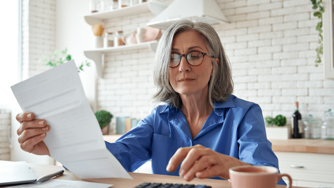 Senior woman with glasses reading IRS debt letter at kitchen table with concerned expression