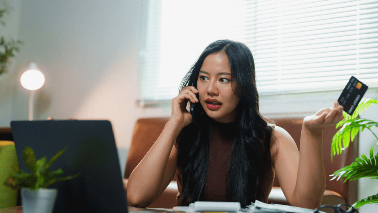 Young woman on phone holding credit card at laptop wondering why do I have a tax debt