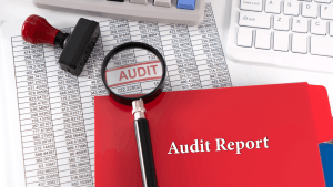 A red Audit Report folder and magnifying glass over financial spreadsheets with an "AUDIT" stamp, alongside a calculator and keyboard on a white desk.