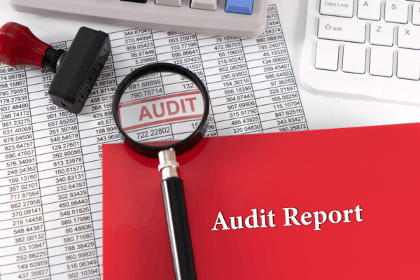 A red Audit Report folder and magnifying glass over financial spreadsheets with an "AUDIT" stamp, alongside a calculator and keyboard on a white desk.