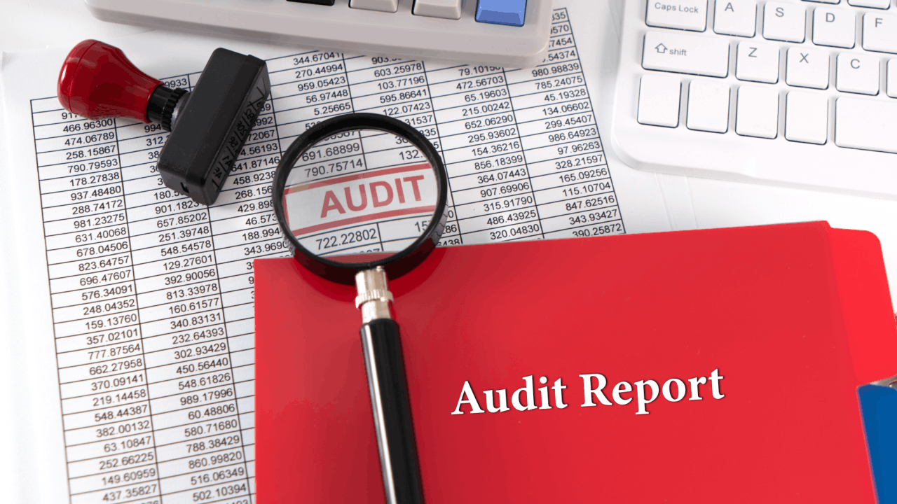 A red Audit Report folder and magnifying glass over financial spreadsheets with an "AUDIT" stamp, alongside a calculator and keyboard on a white desk.
