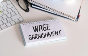 Wage garnishment written on spiral notepad beside keyboard and glasses on white desk