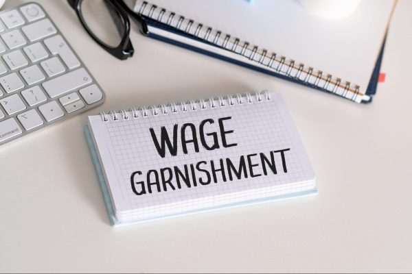 Wage garnishment written on spiral notepad beside keyboard and glasses on white desk